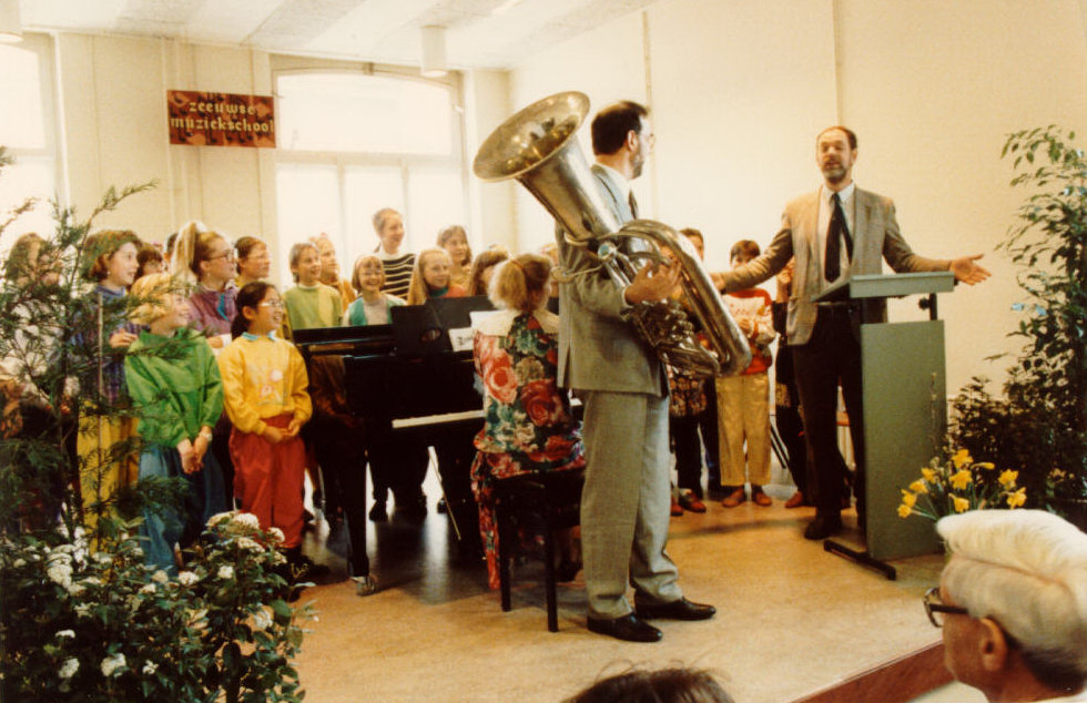 Kinderen staan op een podium bij de piano. Twee mannen staan voor hun, een draagt een tuba