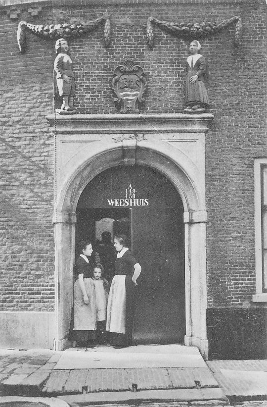Poort van het weeshuis, drie mensen staan in de poort met elkaar te praten. Ze kijken niet in de camera