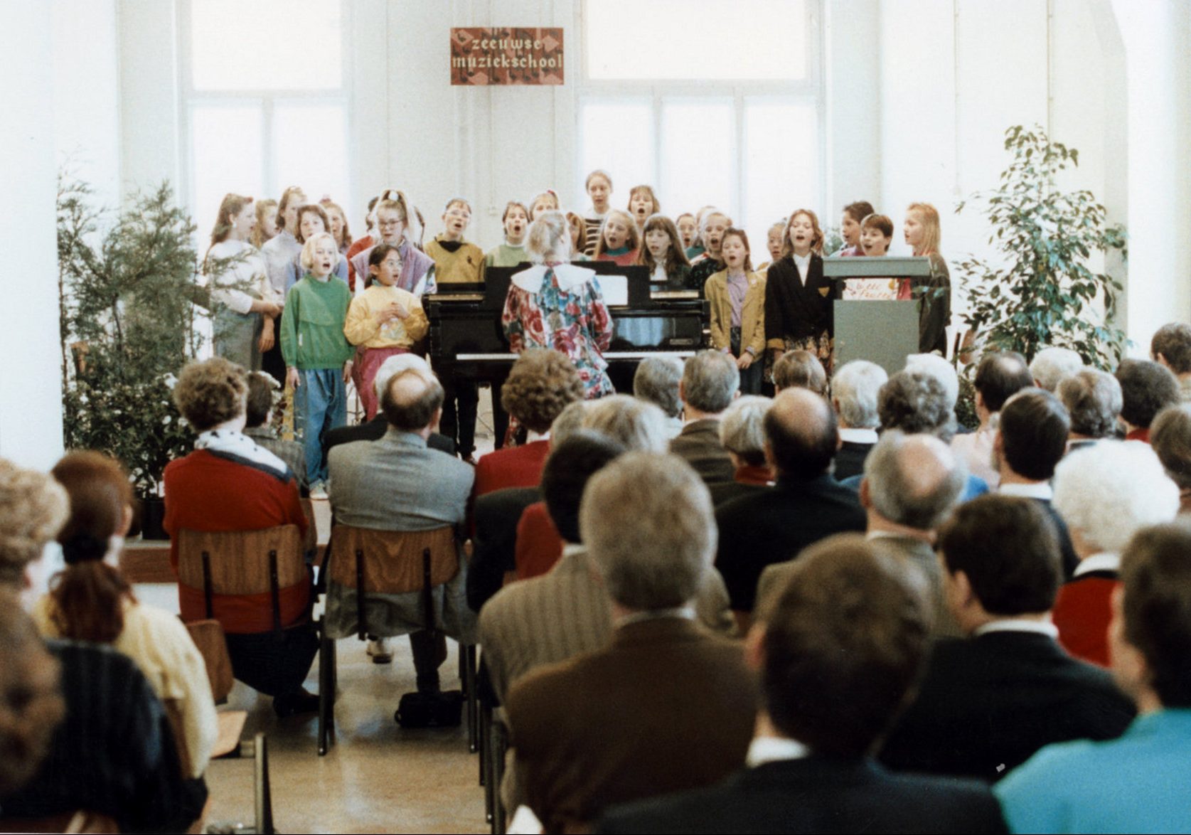 Kinderen zingen op een podium bij de piano, publiek kijkt toe.